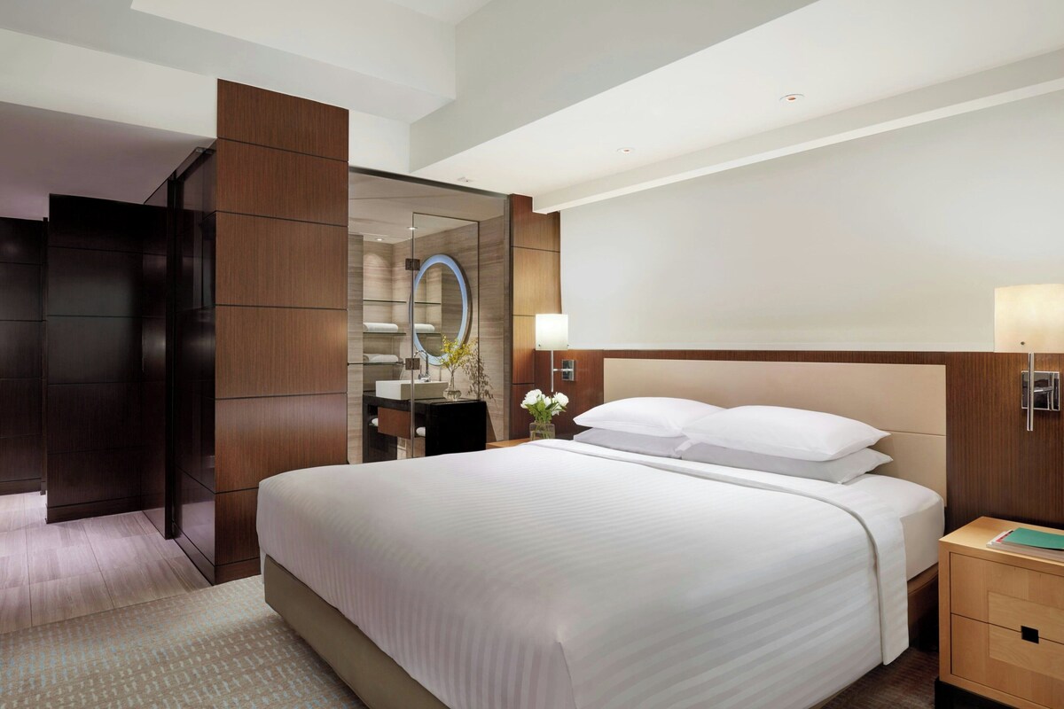 A modern hotel room with a large bed, a wooden wardrobe, a mirror, and a bedside table with a lamp.