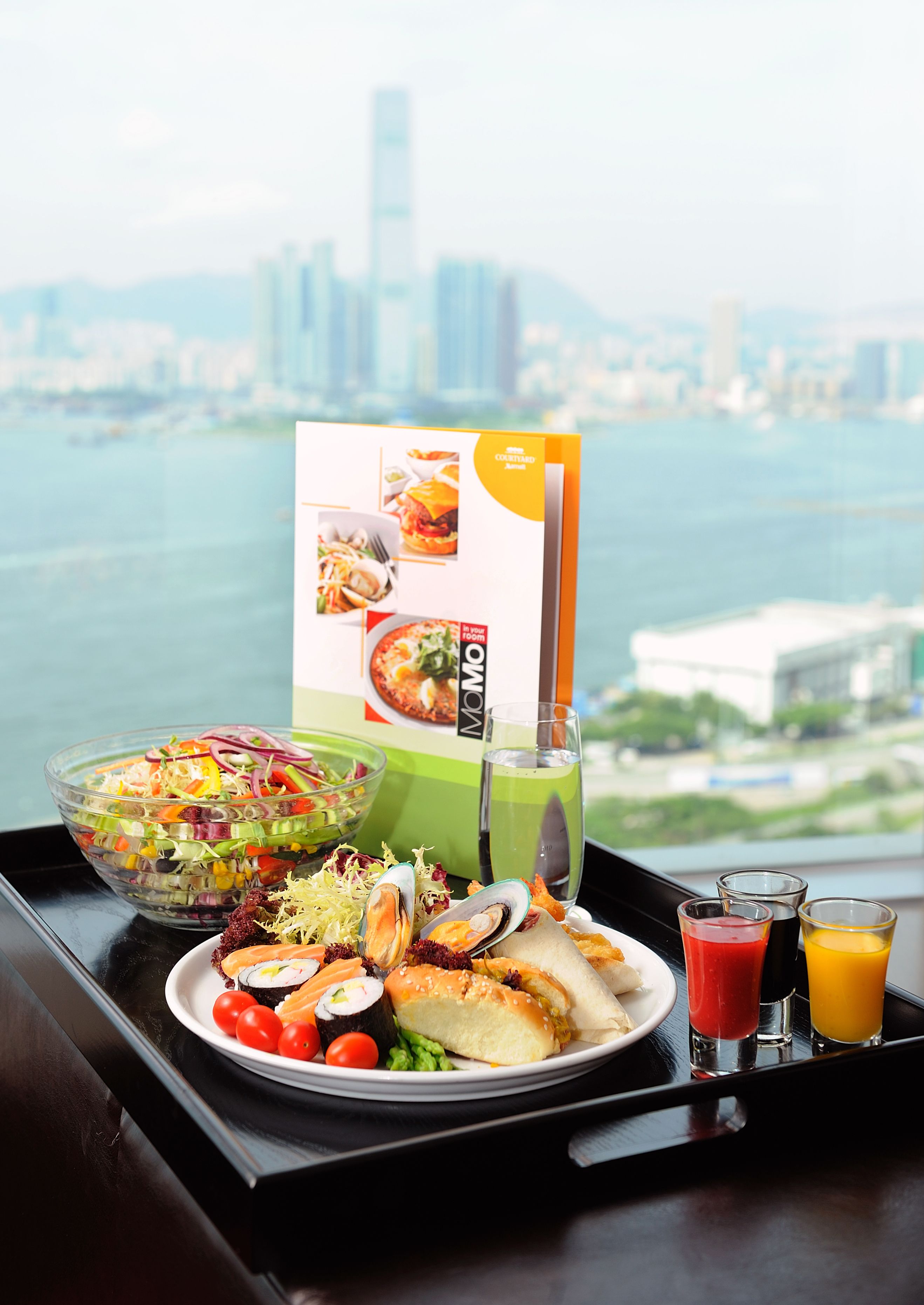 Una bandeja con ensalada, sushi y un menú de bebidas, frente a una vista panorámica de la ciudad.