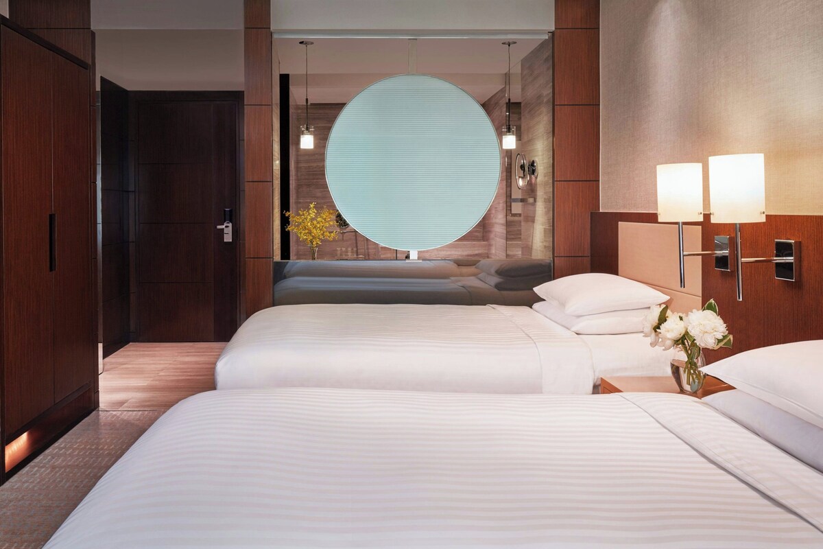 A hotel room with a large circular window, two beds, a nightstand with a vase of flowers, and wall-mounted lamps.