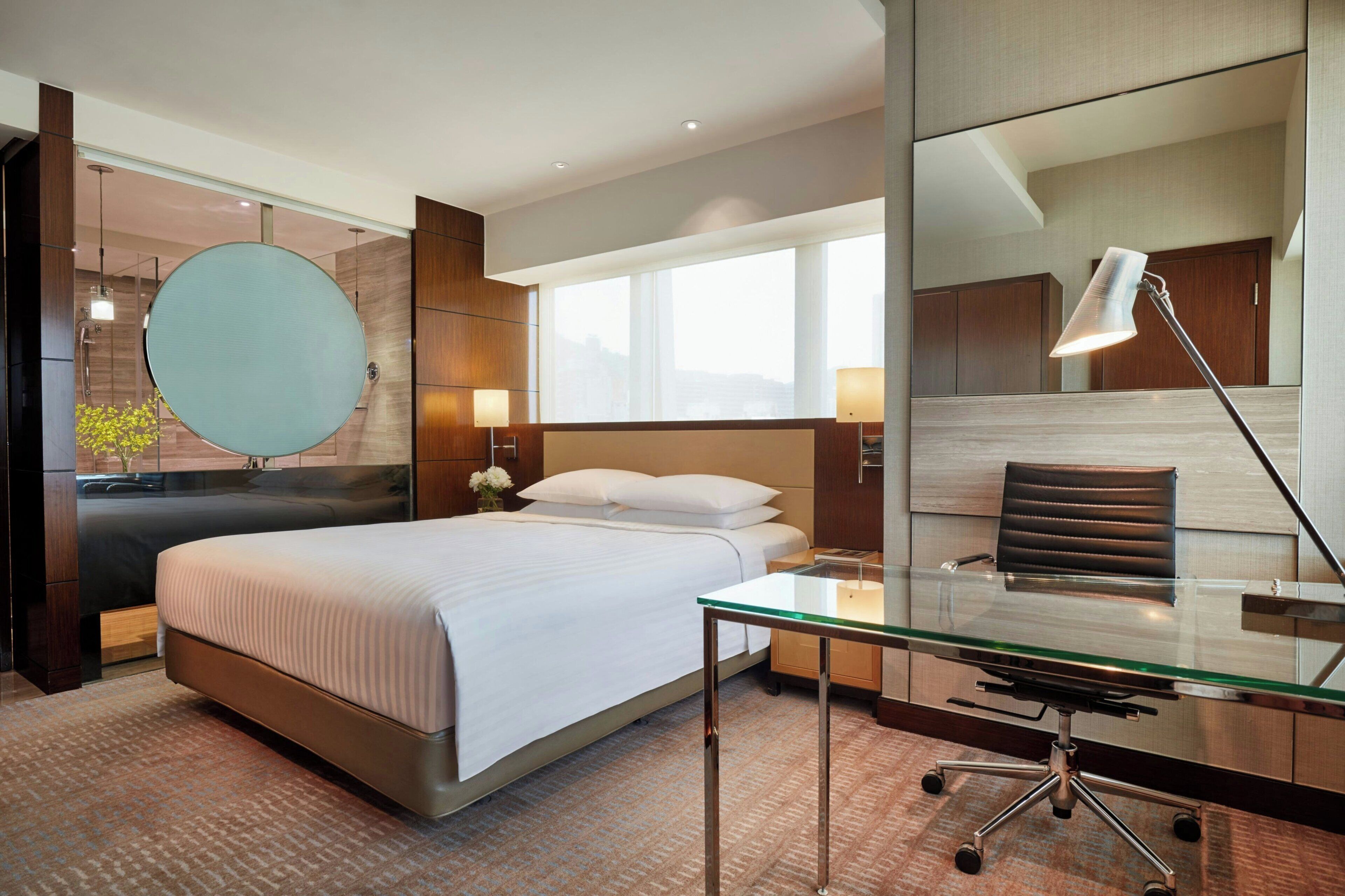 Habitación de hotel con cama, escritorio con silla, espejo redondo y ventana con vistas.