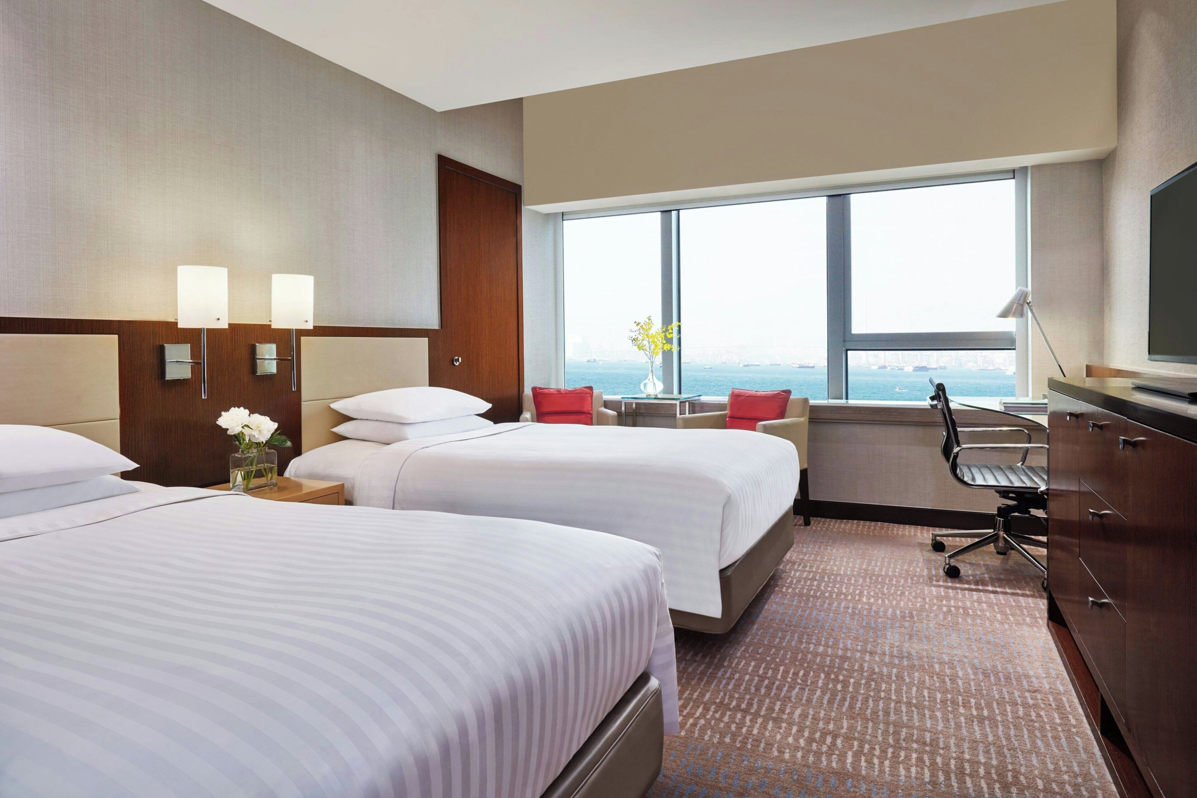 A hotel room with two beds, a desk, a chair, a TV, and a large window with a view.
