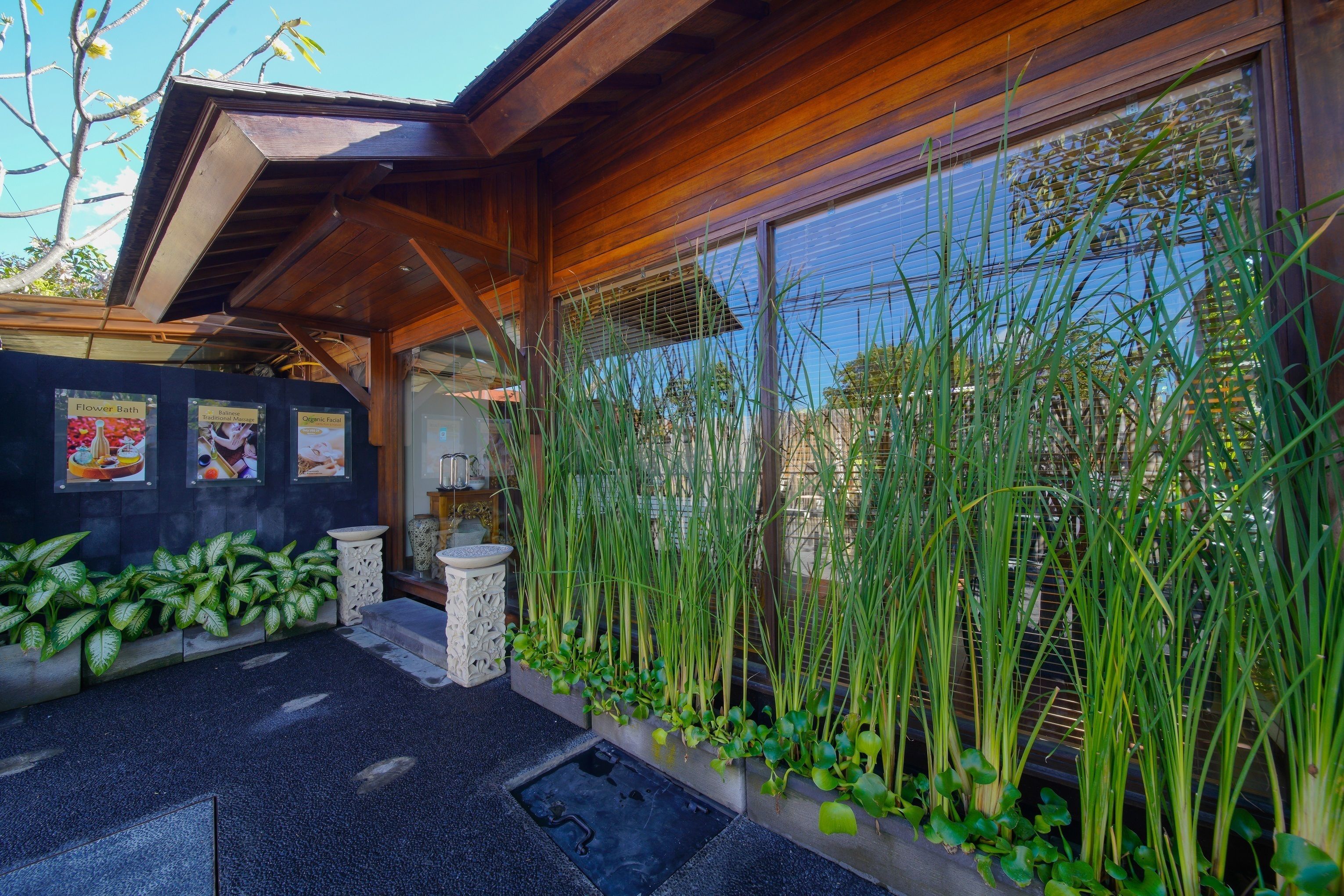 A modern building with a wooden exterior, a black tiled entrance, and a garden area with tall grasses and plants.