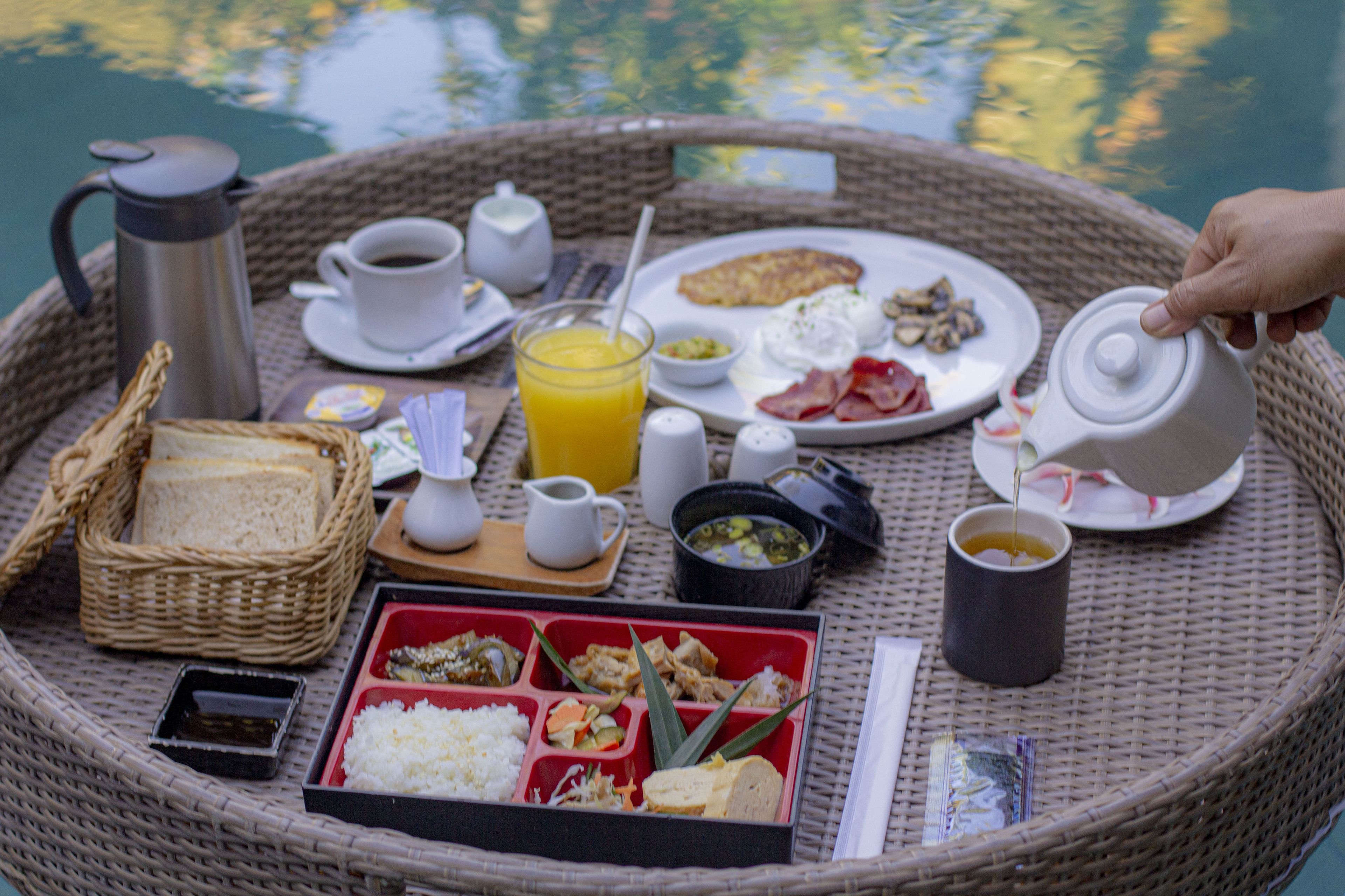 A breakfast tray with various food items and beverages, including eggs, bacon, toast, and a hot beverage, set on a wicker table by a body of water.