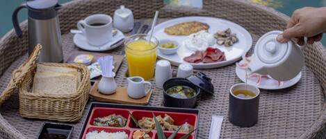 A breakfast tray with various food items and beverages, including eggs, bacon, toast, and a hot beverage, set on a wicker table by a body of water.