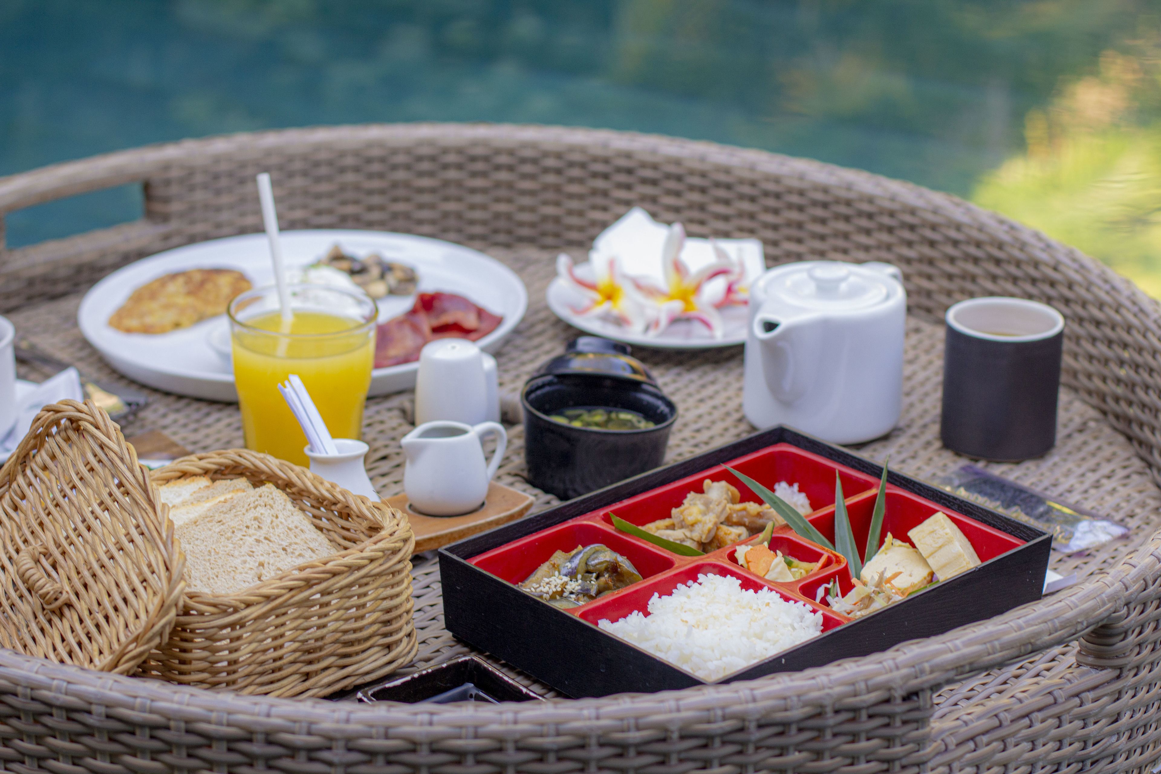 Une table en osier, dressée pour un repas, avec des assiettes, une boisson et une boîte contenant du riz et des légumes.