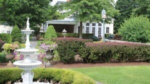 A garden with a fountain, neatly trimmed hedges, and a building with a covered entrance. - The Bykenhulle House (Hopewell Junction)