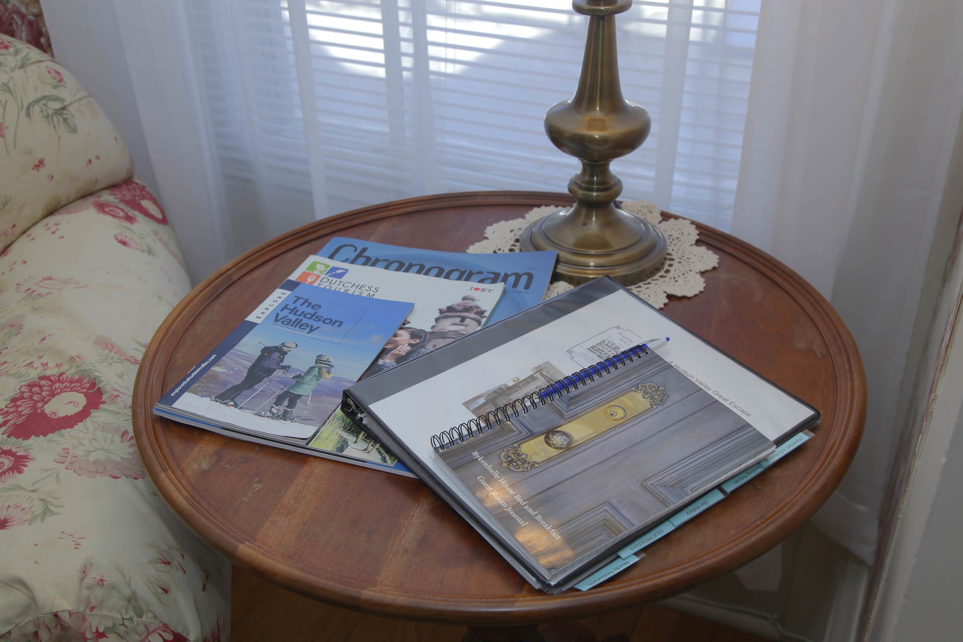 Une petite table d’appoint ronde en bois, sur laquelle se trouvent une lampe en laiton et quelques brochures de voyage.