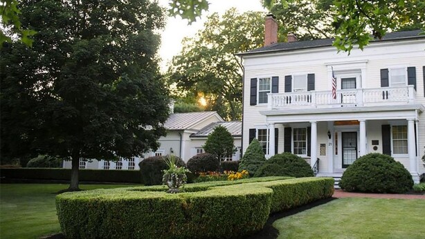 A two-story white house with a balcony, surrounded by well-manicured hedges and trees. - The Bykenhulle House (Hopewell Junction)