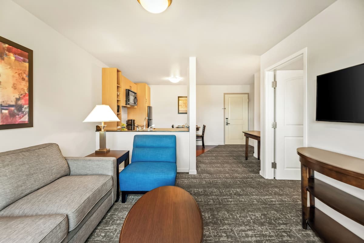 A hotel room with a grey sofa, a blue armchair, a wooden coffee table, a TV mounted on the wall, a kitchen area with a microwave, and a dining table.