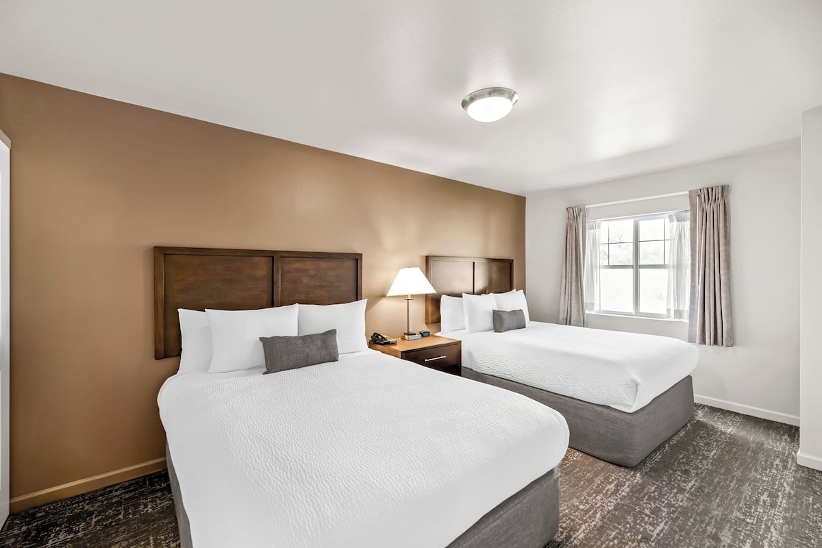 A hotel room with two beds, a nightstand with a lamp, a window with curtains, and a ceiling light.