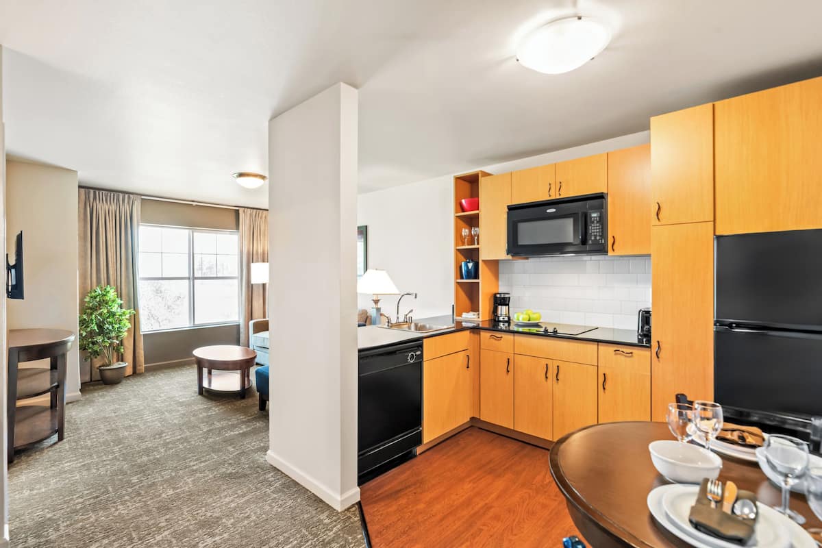 A modern kitchen with wooden cabinets, a black microwave, and a black refrigerator. There is a dining table with plates and glasses, and a small round table with a lamp.