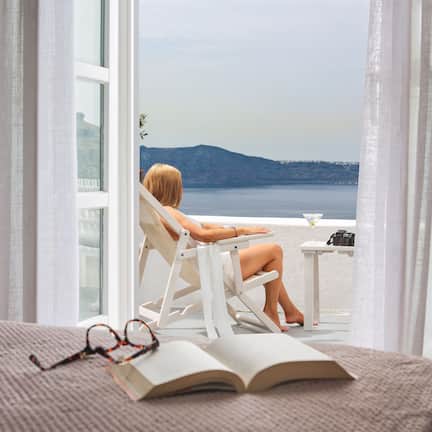 A room with a bed, an open book, and a pair of glasses on the bed. There is a window with curtains, through which a person is sitting on a chair, overlooking a body of water and a distant mountain.