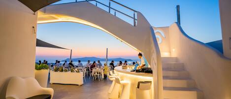 A modern outdoor seating area with a bar, white furniture, and a view of the sea at dusk.
