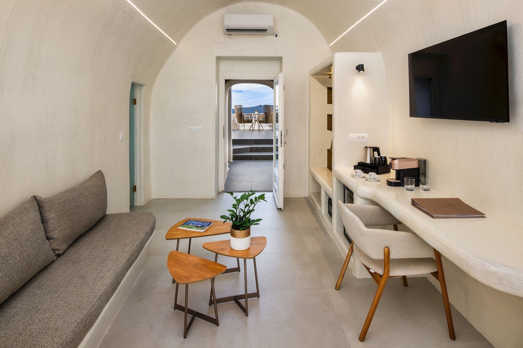 A modern interior with a sofa, a small dining area with stools, a table with a plant, and a TV mounted on the wall.