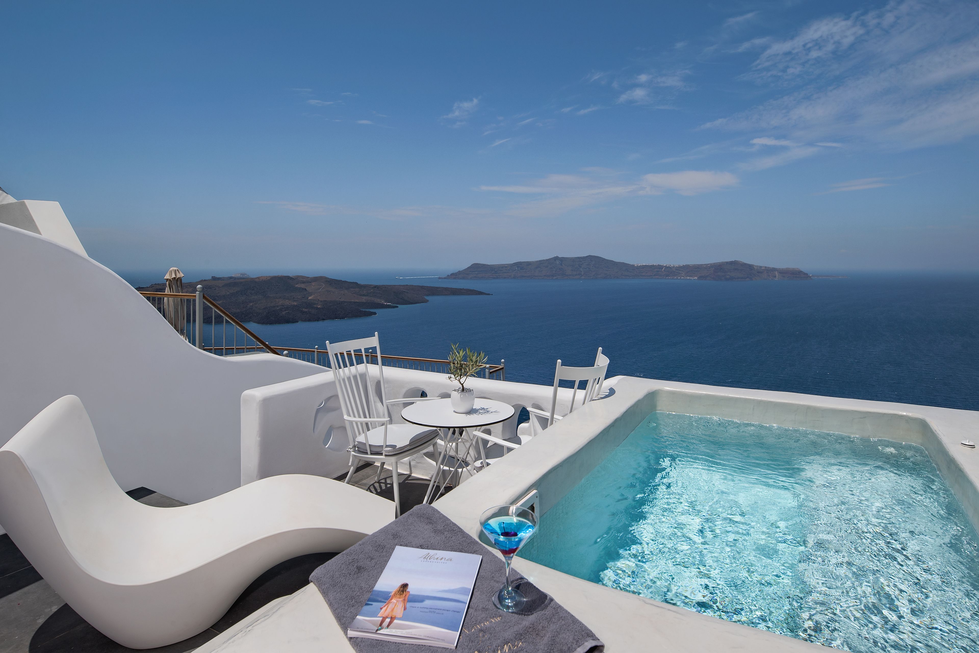 Zona de piscina con vista al mar, sillas reclinables blancas, una mesa y una copa de martini.