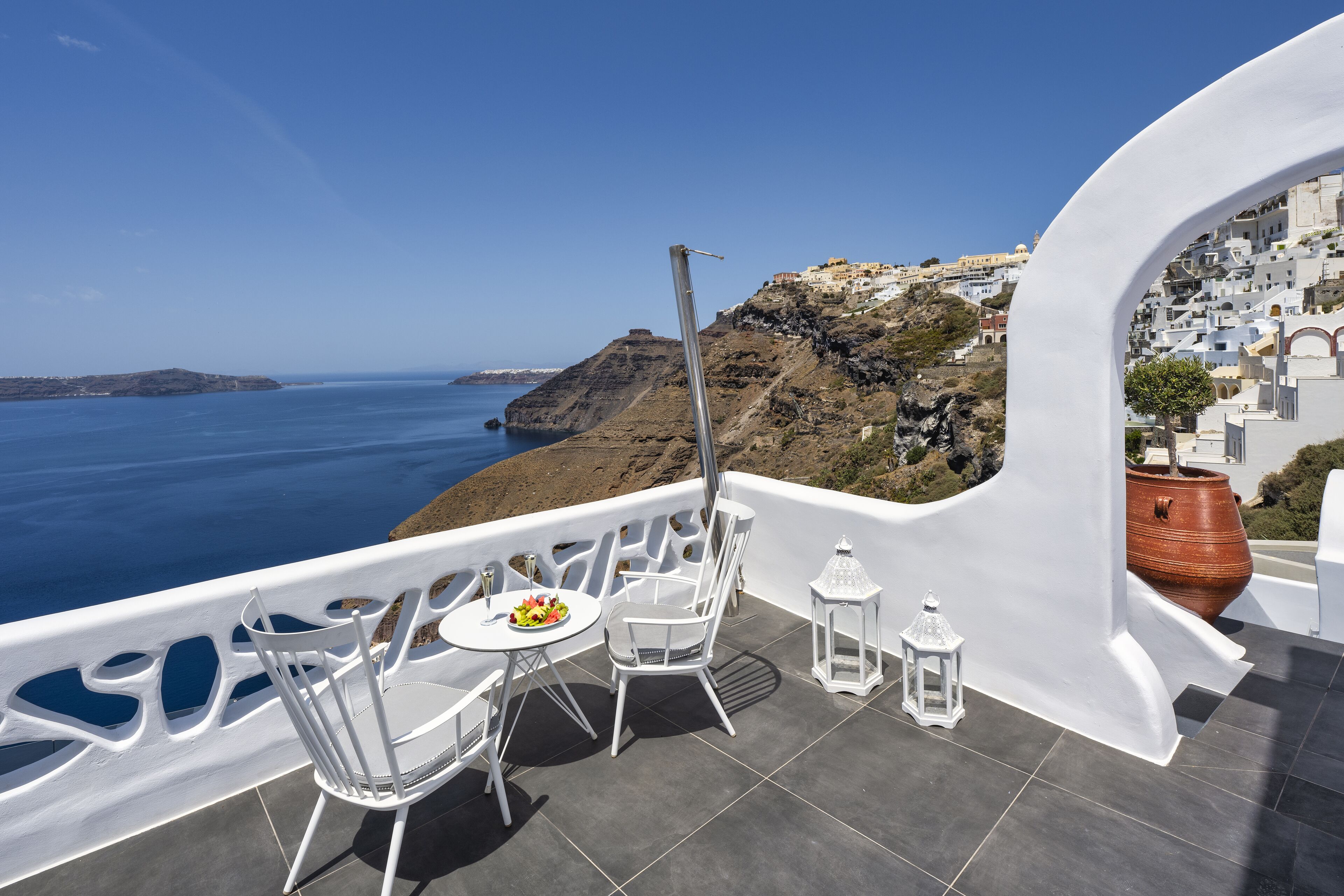 A balcony with a table, chairs, and a potted plant, overlooking a coastal town and the sea.