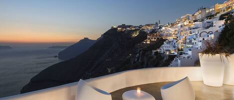 A seaside balcony with white, modern seating and a small table with a candle, overlooking a coastal town at dusk.