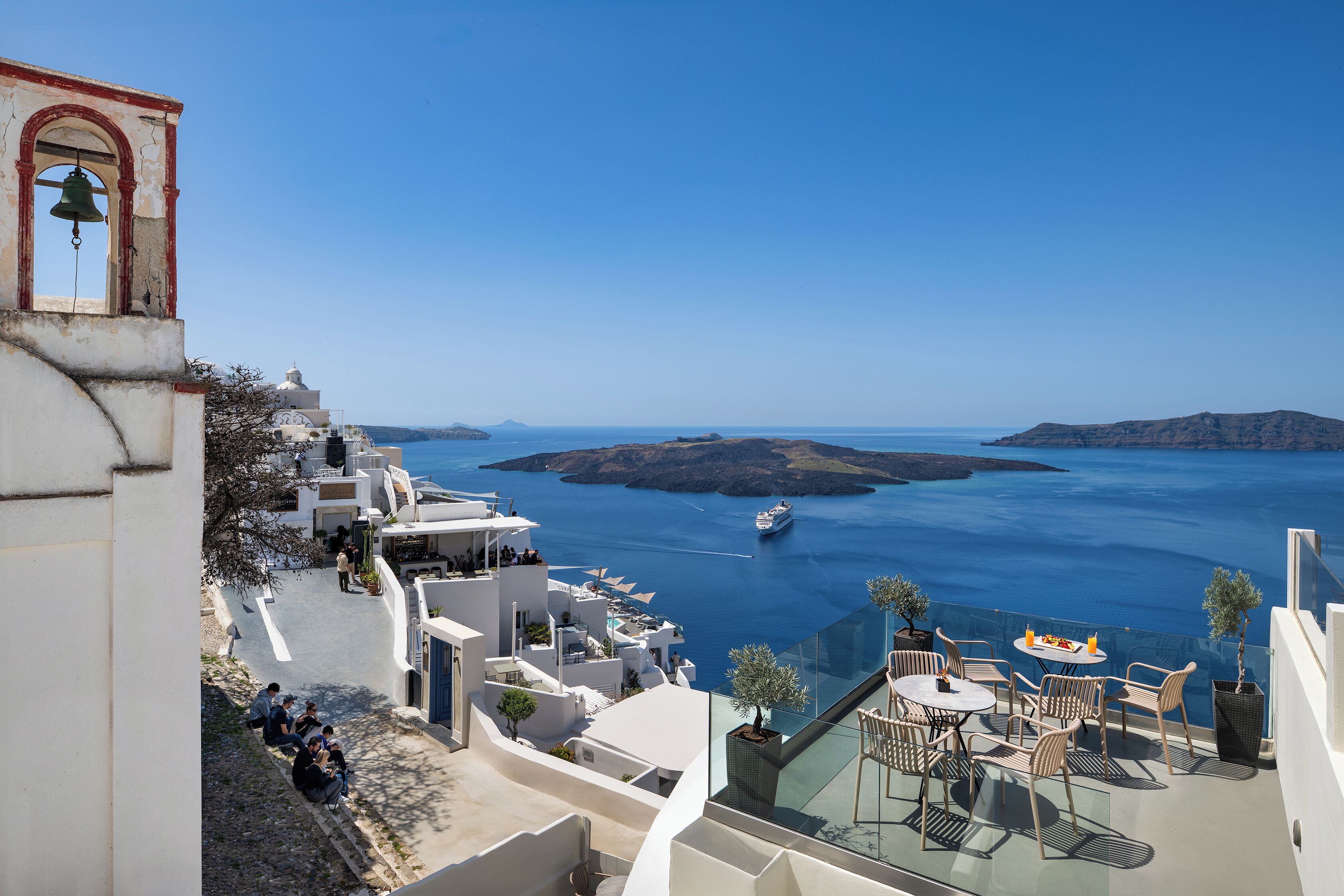 Un hotel costero con terraza con vistas al mar, una torre de campanas y un comedor con mesas al aire libre.