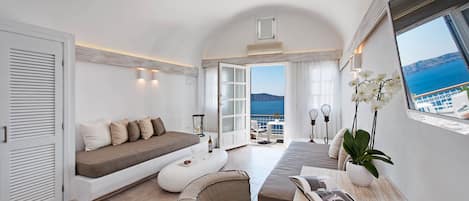 A modern, well-lit room with a large window offering a view of the sea. It includes a sofa, a small table with a bottle, and a potted plant.