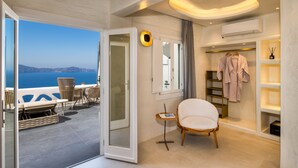 Habitación de hotel moderna con balcón que da al mar, zona de estar con mesa y sillas, sillón blanco y vestidor con batas colgadas.