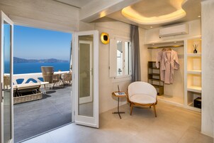 A modern hotel room with a balcony overlooking the sea, a seating area with a table and chairs, a white armchair, and a walk-in closet with hanging robes.