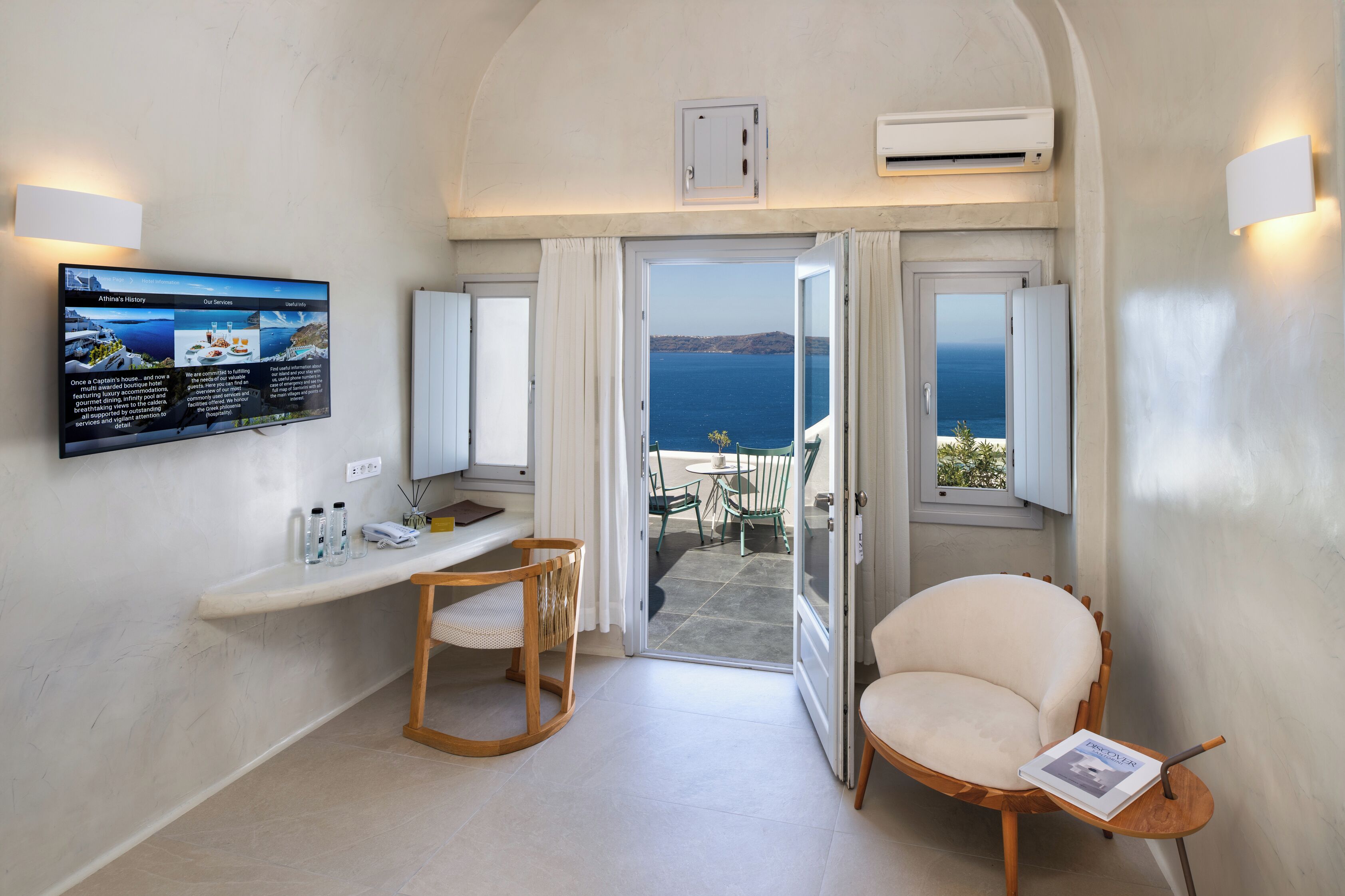 Een kamer met een tv waarop een menu staat, een houten stoel, een klein tafeltje en uitzicht op zee door een geopende deur.