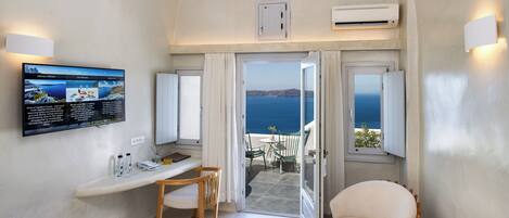 Une pièce avec un téléviseur affichant un menu, une chaise en bois, une petite table et une vue sur la mer par une porte ouverte.