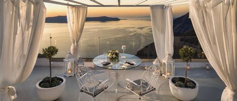 A terrace with a glass table, white chairs, and potted plants, overlooking a scenic landscape.