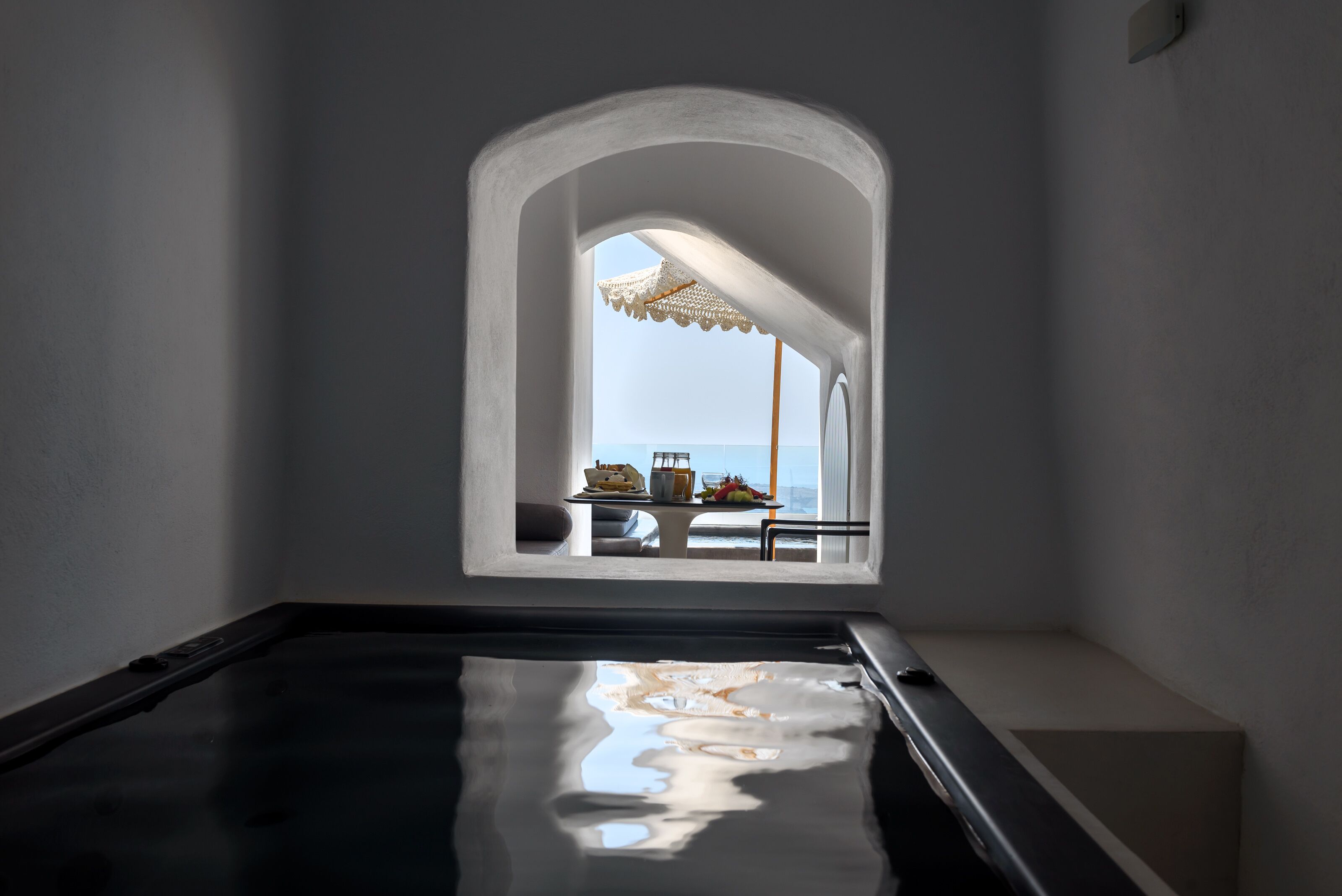 Quarto com vista para o mar por uma janela em arco, piscina e área de jantar com mesa e cadeiras.