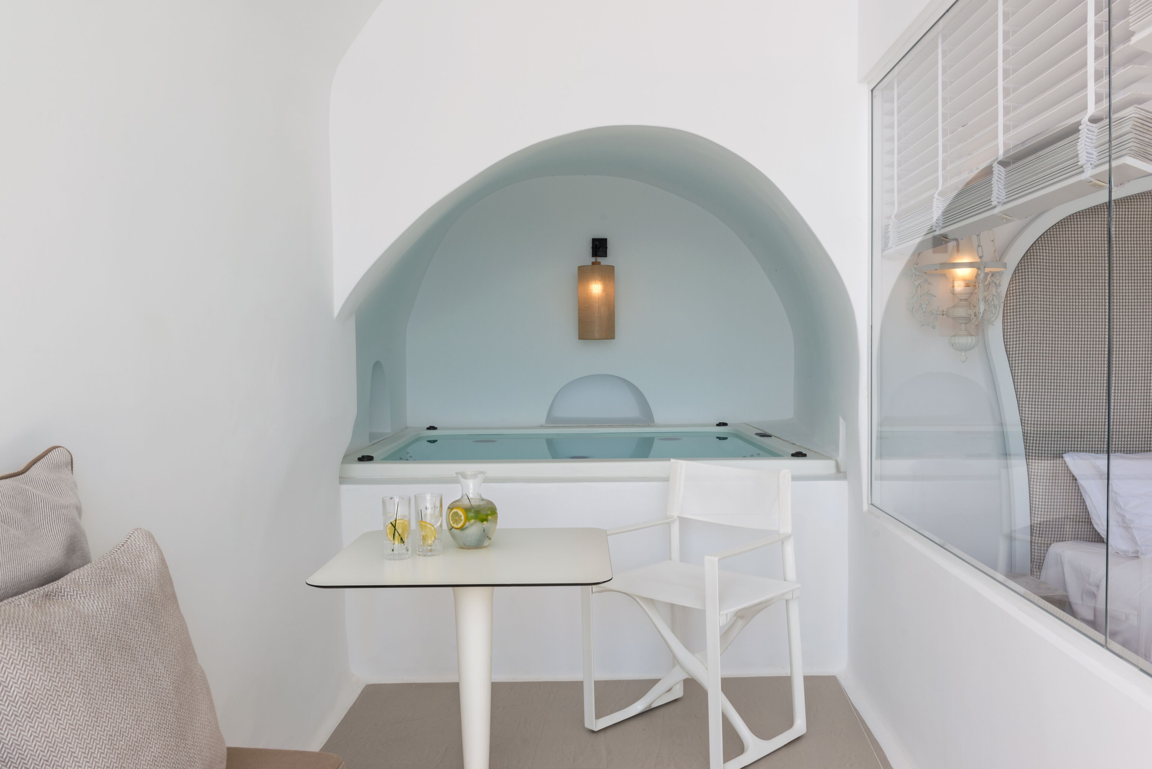 A small, white room with a glass-top table, a white chair, a white sofa, and a window with white blinds.