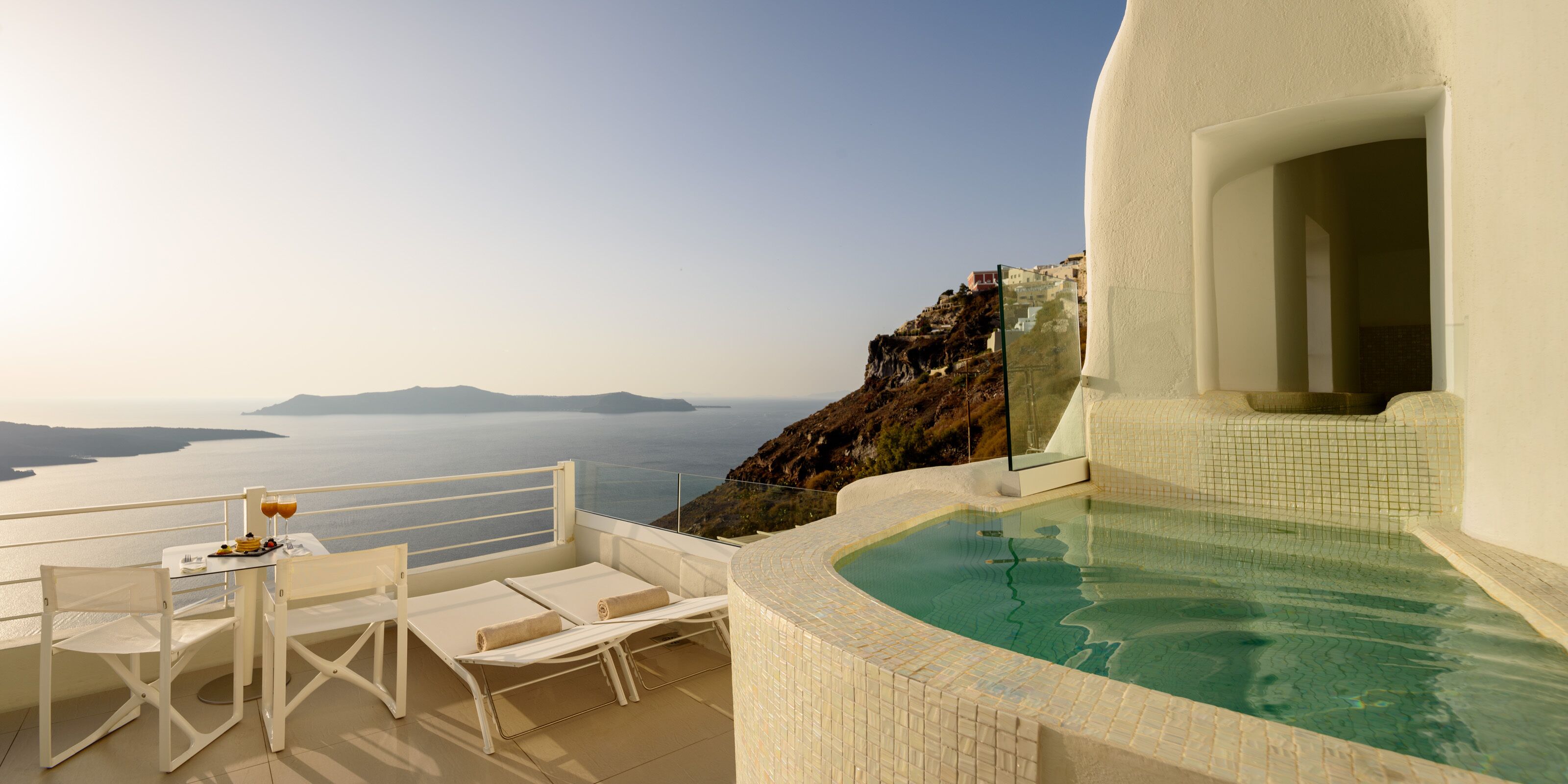 A sunlit terrace with a pool, white furniture, and a view of the sea and distant islands.