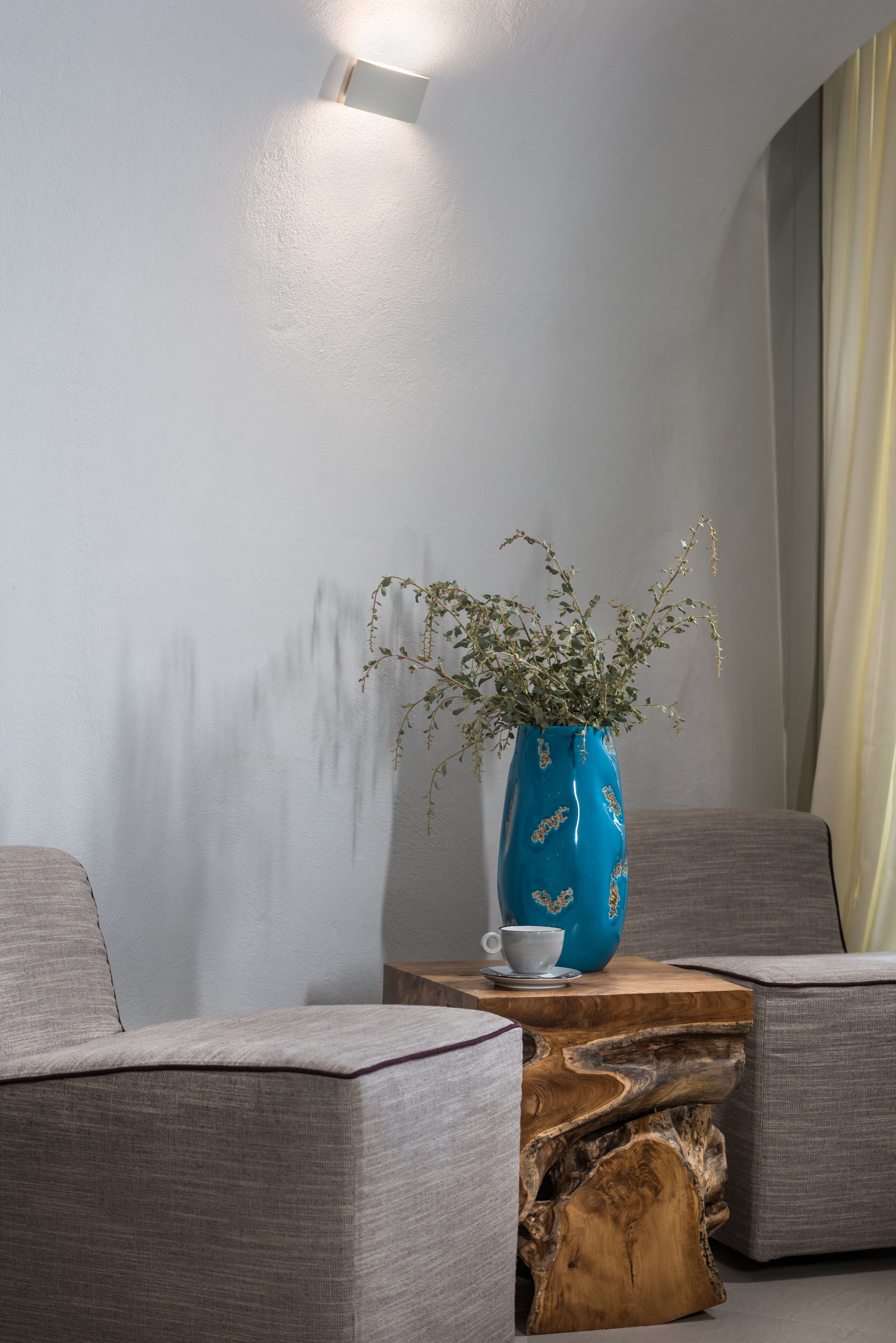 A modern living room with a blue vase on a wooden side table, grey sofas, and a white wall with a light fixture.