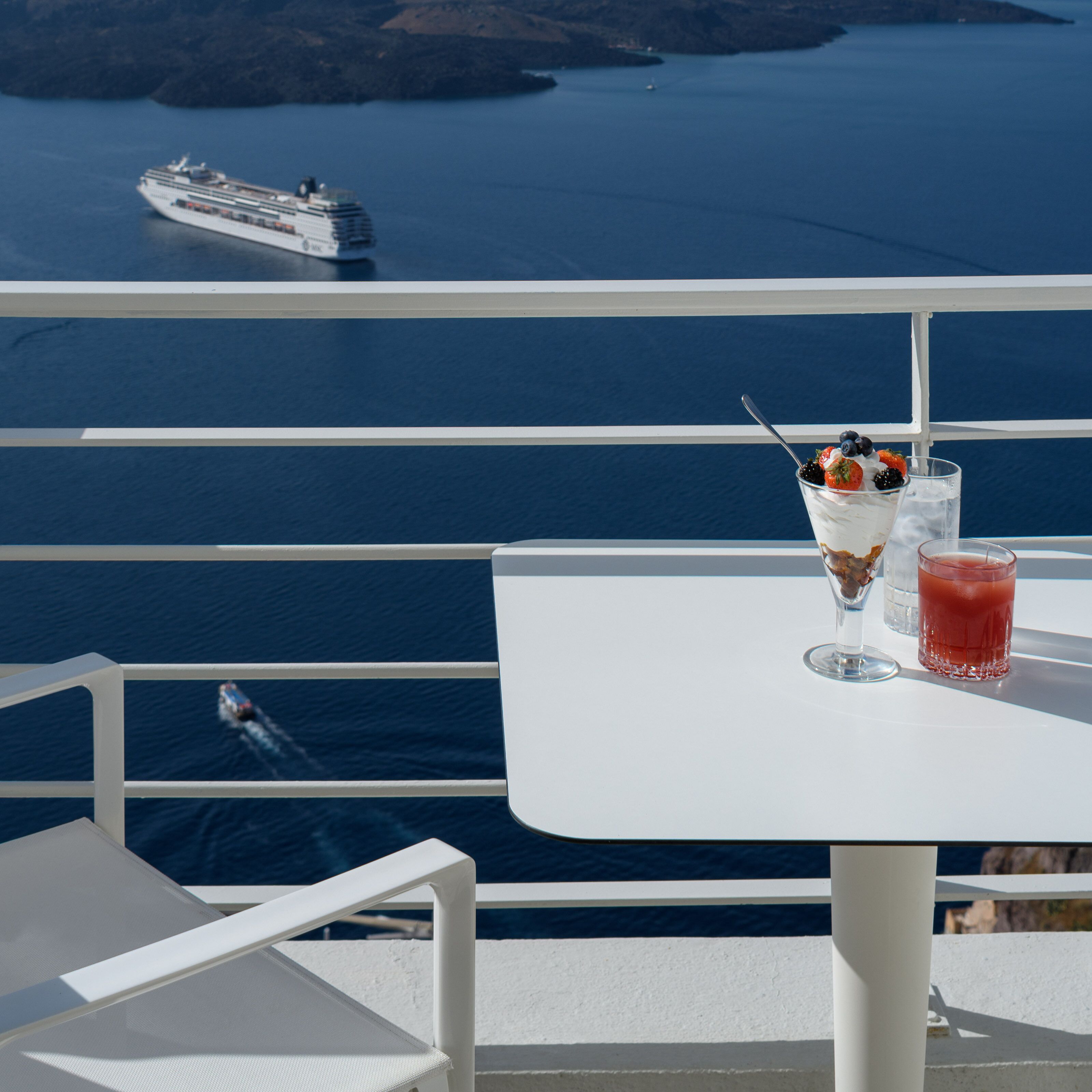 A balcony with a table, two drinks, and a view of a cruise ship on the water.