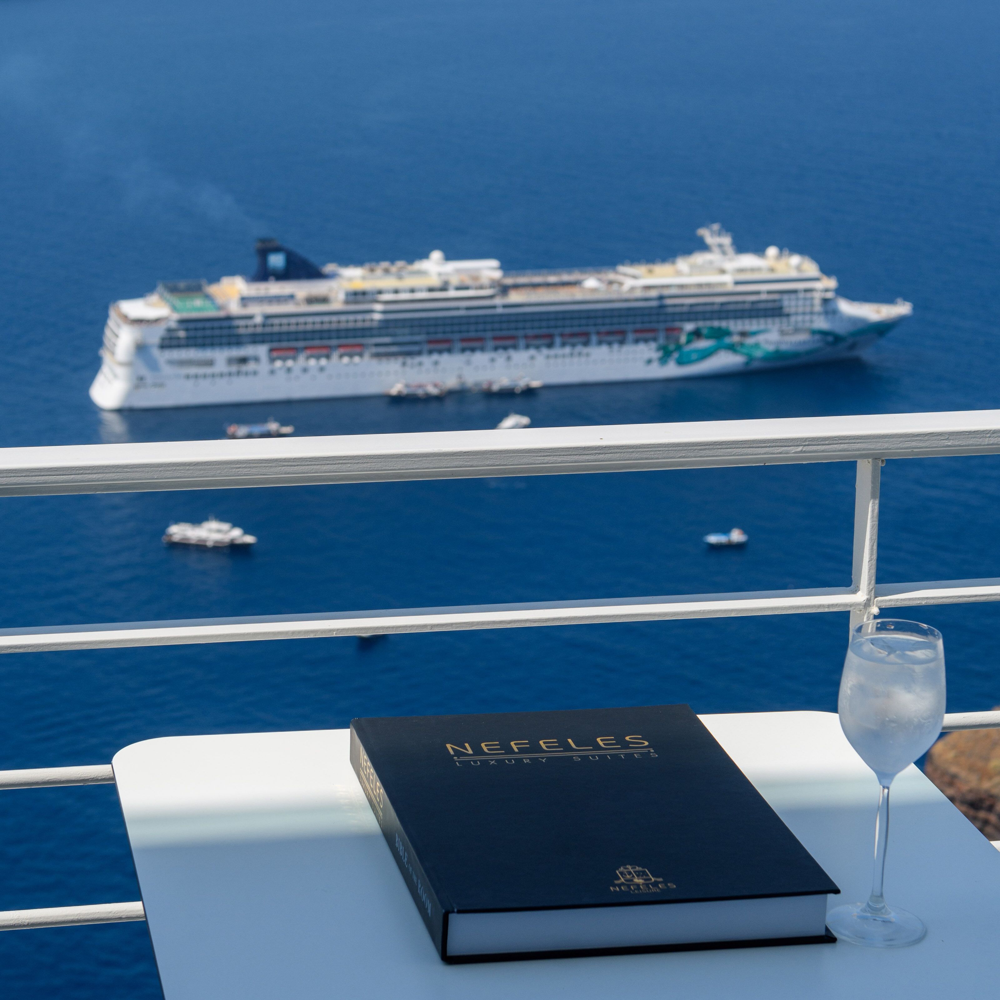 A book titled "NEFFLEYS LUXURY SUITES" on a white table with a glass of water beside it, overlooking a cruise ship on the water.
