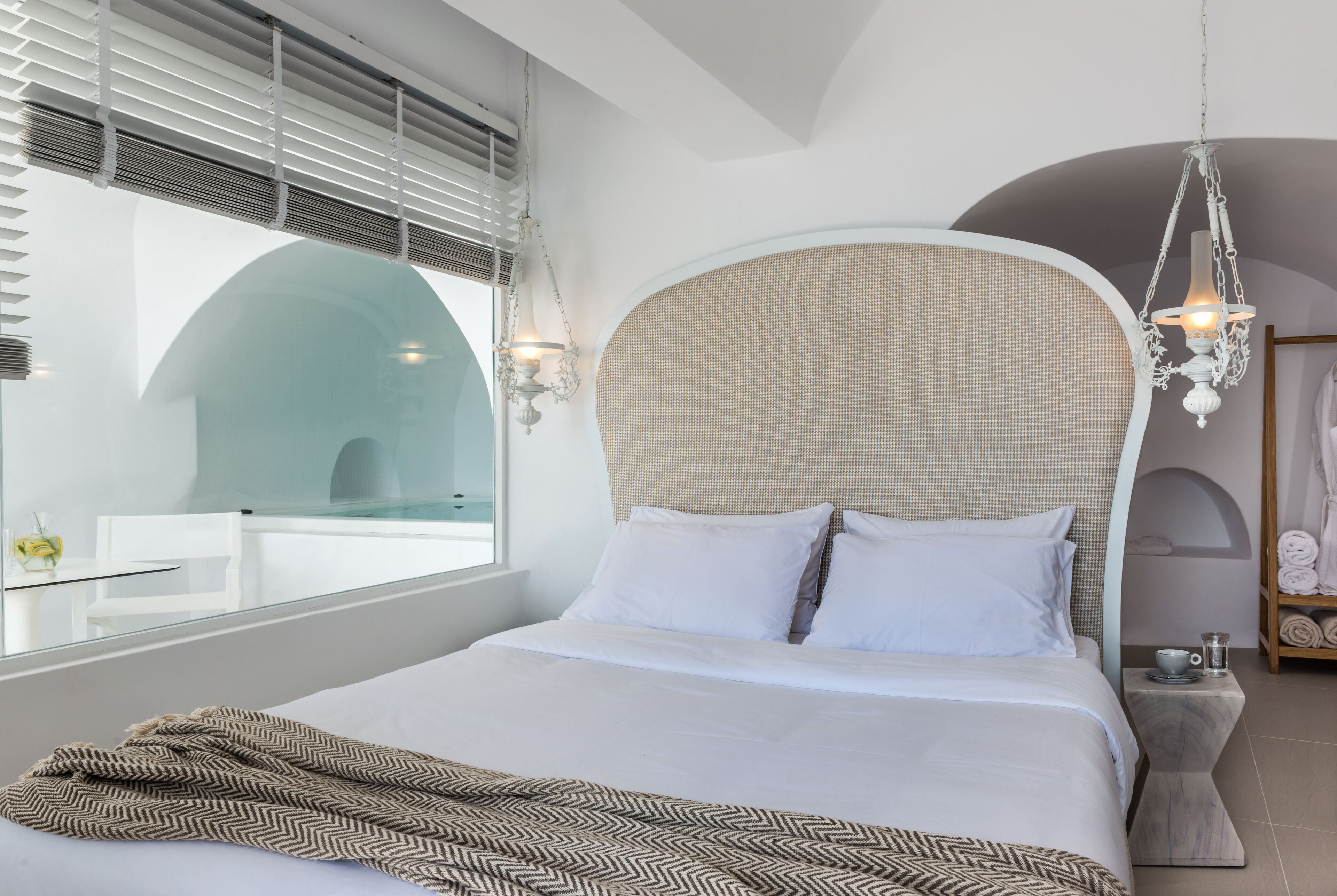 A modern bedroom with a large bed, white bedding, a patterned throw, a hanging light fixture, and a view of a glass-walled room.