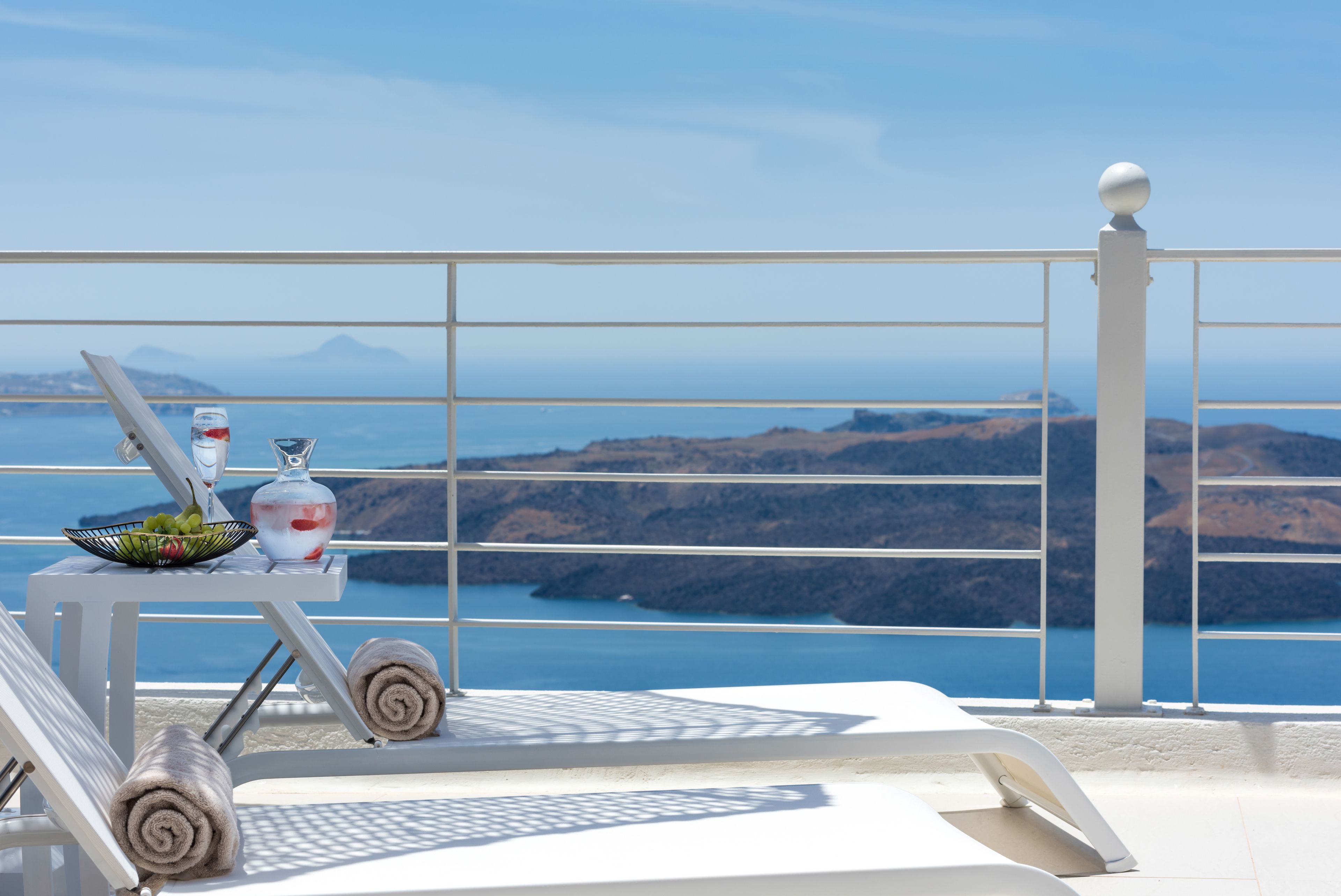A balcony with a white railing, a table with a glass vase, a bowl of fruit, and rolled towels.
