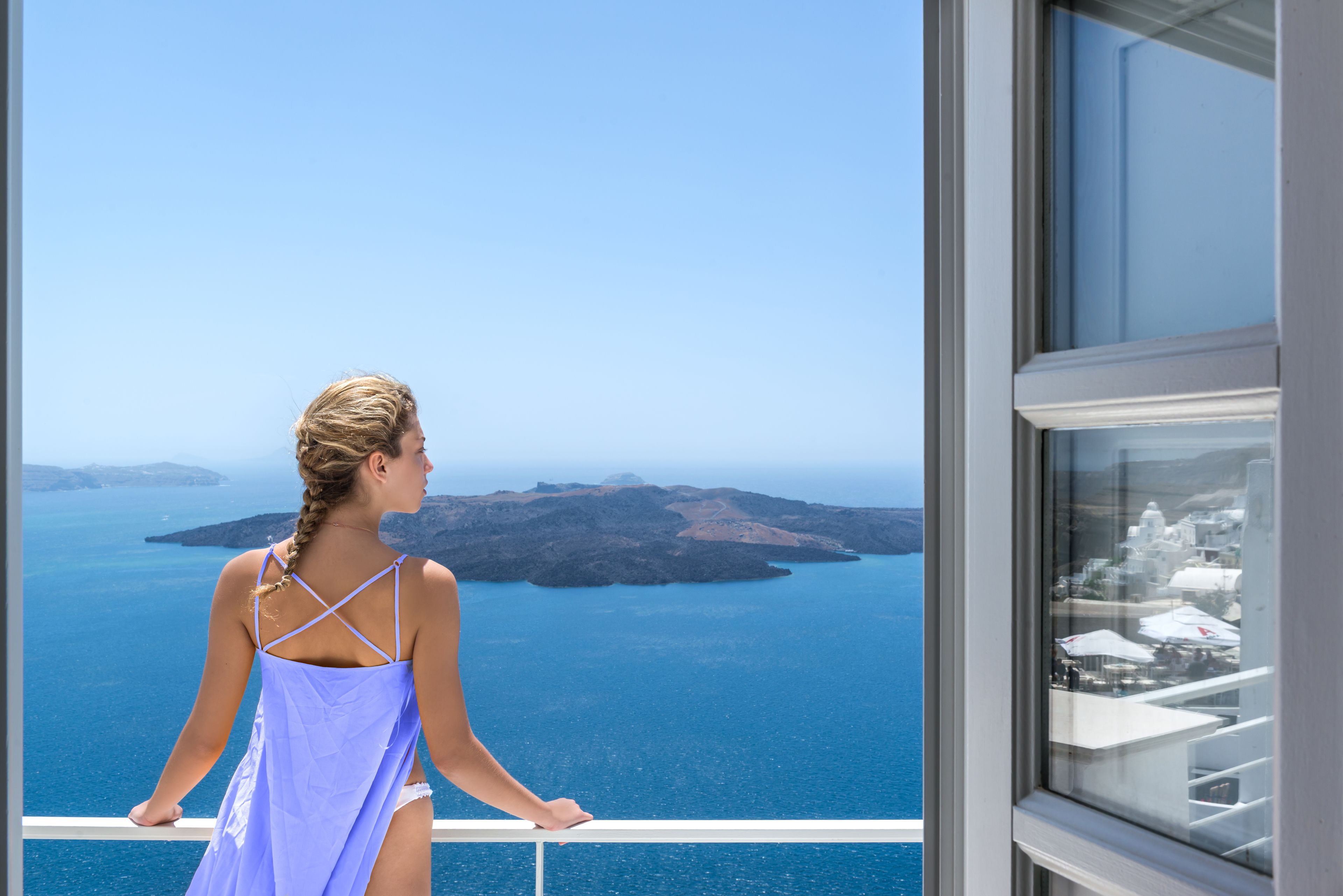 A woman in a purple dress standing by a balcony with a view of the sea and islands.