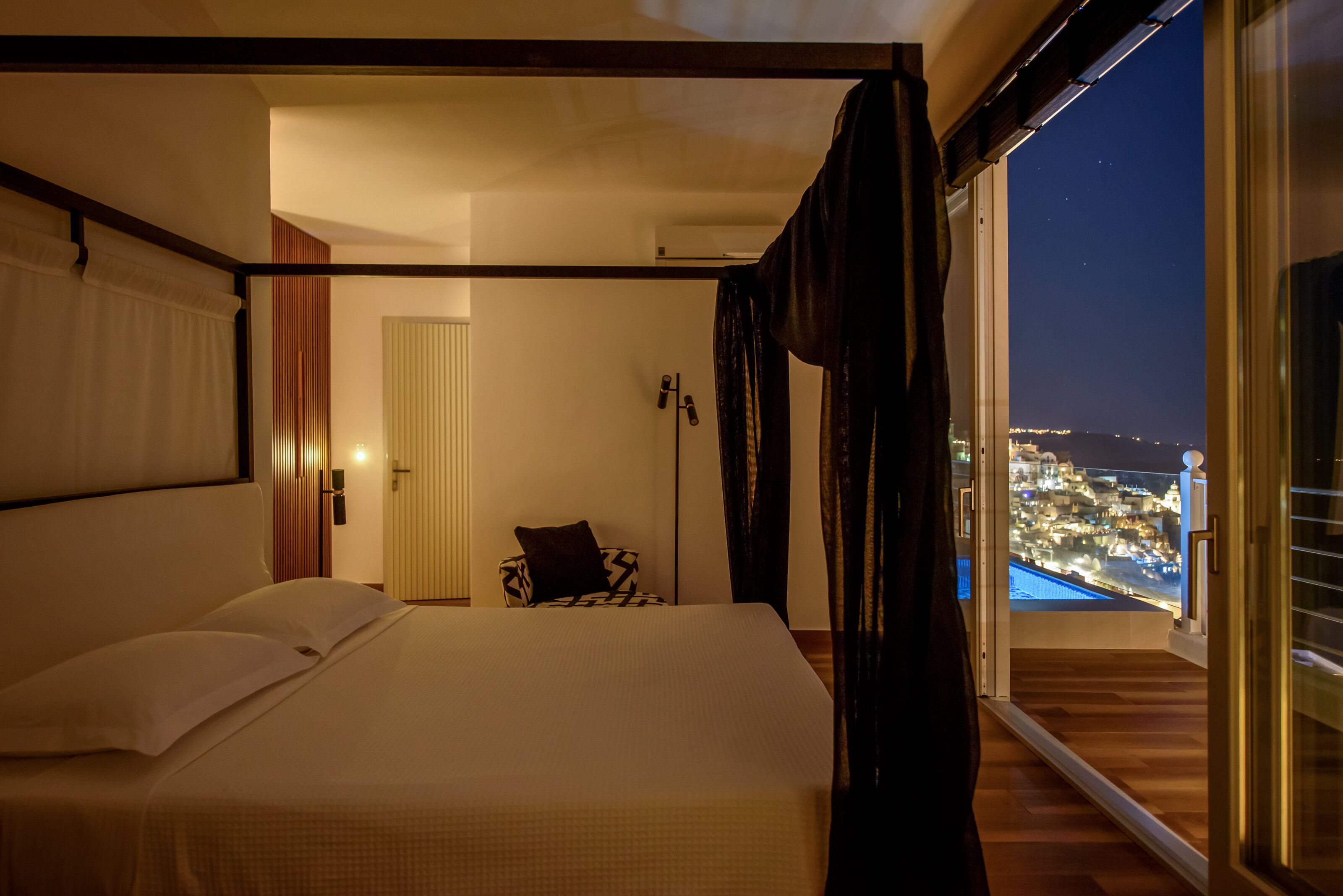 A bedroom with a large bed, a view of a cityscape through a balcony, and a door leading to another room.