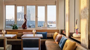 A cozy seating area with a sofa and chairs, a table with a vase and books, and a view of a cityscape through large windows.