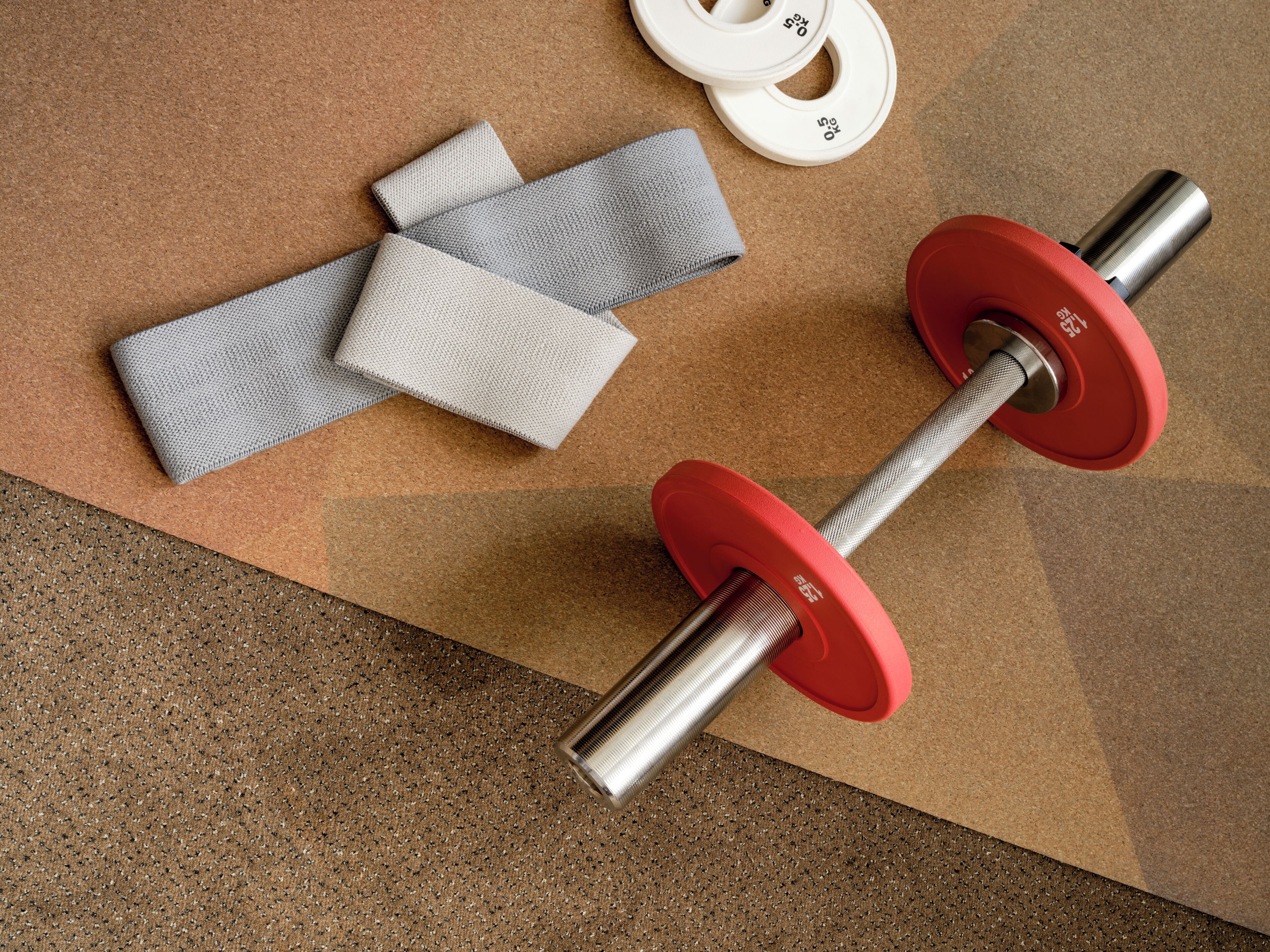 Equipamento de musculação com barra, pesos vermelhos e munhequeiras cinzas, tudo sobre um fundo de cortiça.