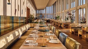 A spacious restaurant with long tables and booths, large windows, and a high ceiling.