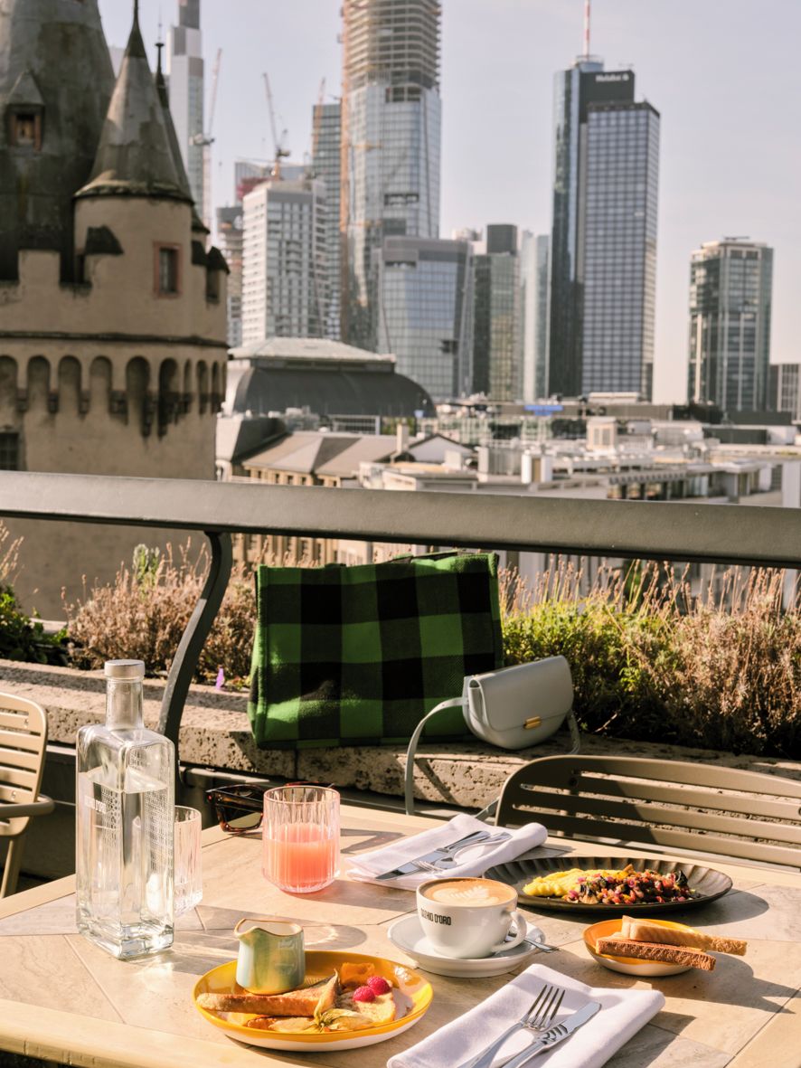 A table with breakfast items, a glass of juice, and a bottle of water, overlooking a cityscape with historic and modern buildings.