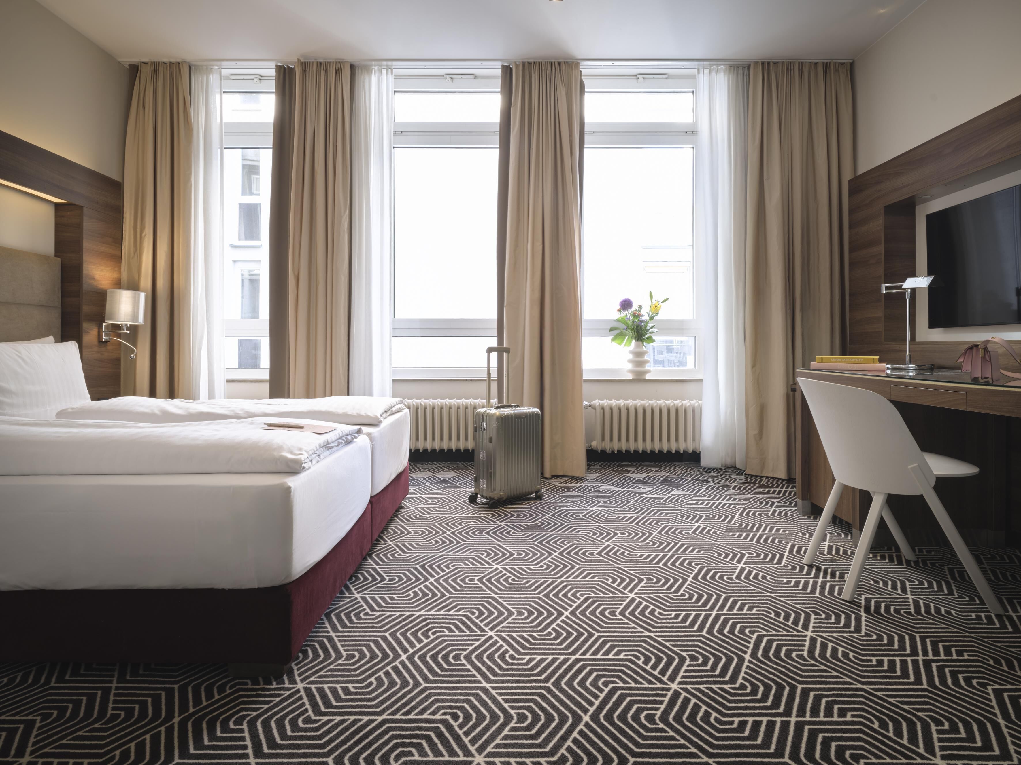 Une chambre d’hôtel avec un grand lit, un bureau avec une chaise, une valise et une fenêtre avec des rideaux.