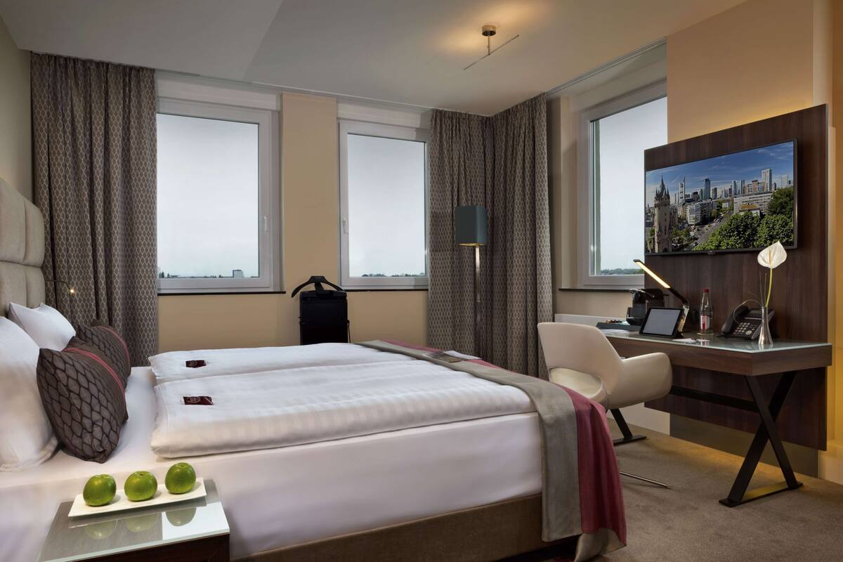 Ett hotellrum med en stor säng, ett skrivbord och en stol. Det finns gardiner, en lampa och en tavla på väggen.