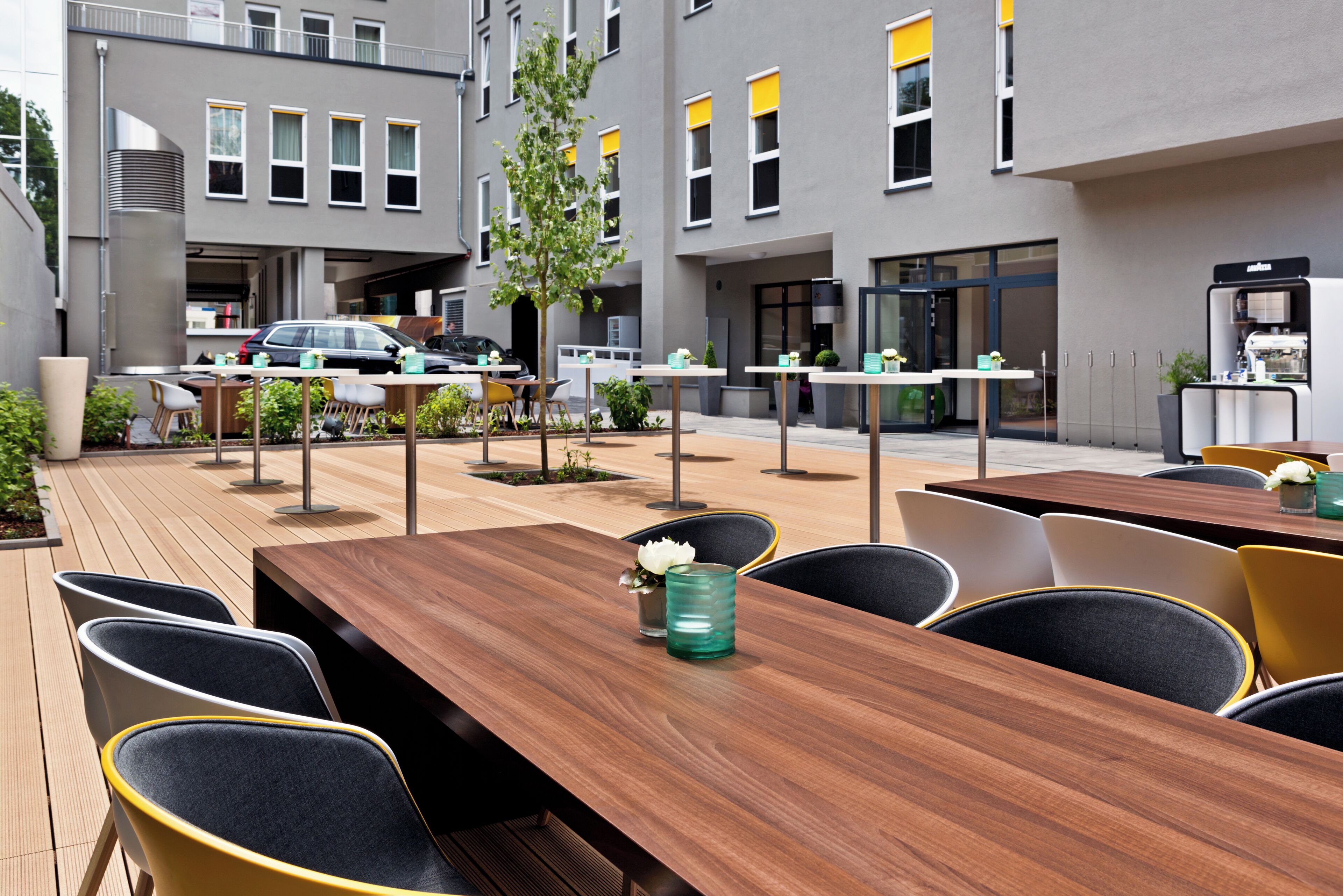 Un espace de restauration en plein air, avec des tables et des chaises en bois, entouré d’un bâtiment moderne doté de grandes fenêtres et où une voiture est garée à proximité.