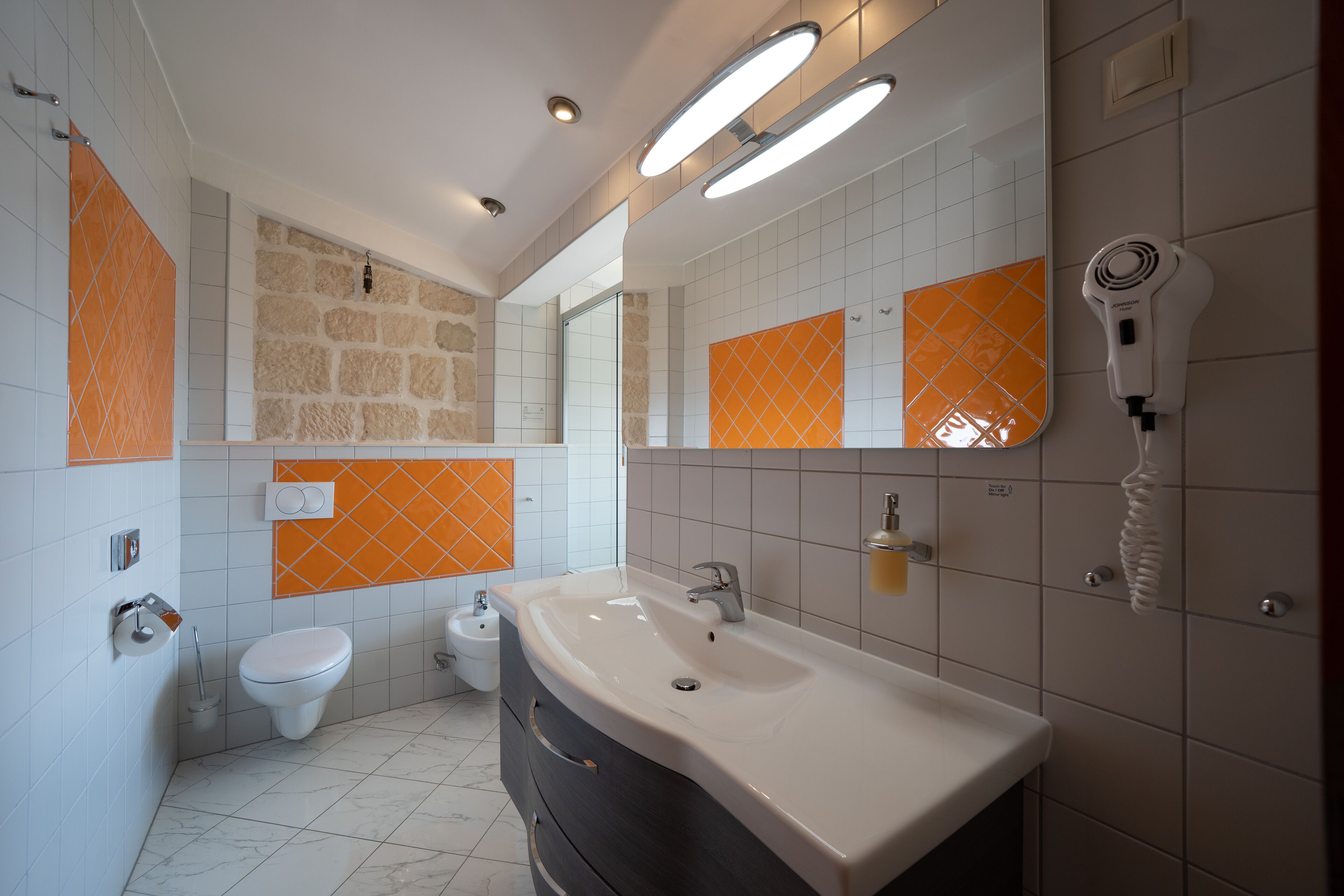 A modern bathroom with a large sink, a toilet, and a bidet. There are orange and white tiles on the walls, and a hairdryer mounted on the wall.
