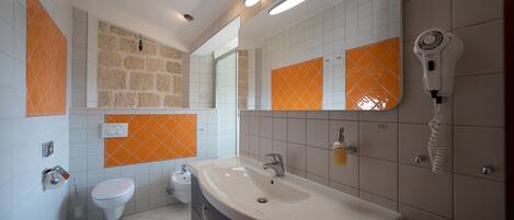 A modern bathroom with a large sink, a toilet, and a bidet. There are orange and white tiles on the walls, and a hairdryer mounted on the wall.