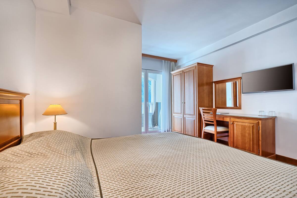 Una habitación de hotel con cama, escritorio, silla, televisor y ventana con cortinas.
