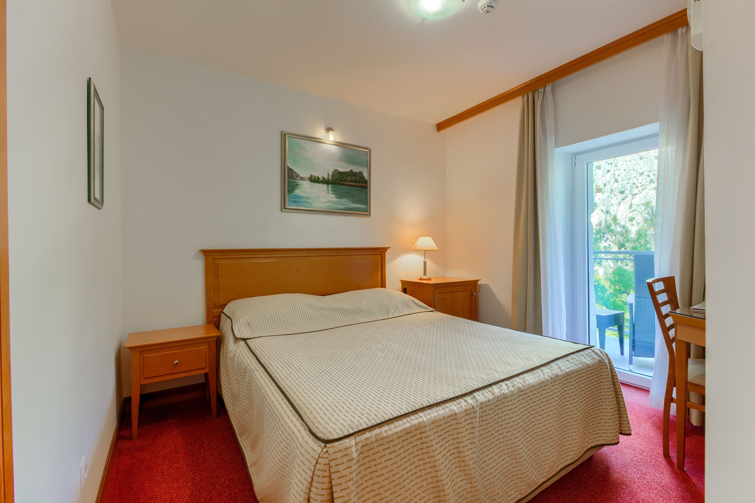 Une chambre à coucher comprenant un lit, une table de chevet, une lampe, une chaise et un balcon donnant sur un espace verdoyant.