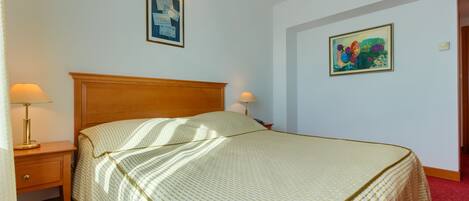 A neatly made bed with a patterned bedspread, a wooden headboard, and two bedside lamps. There are two framed pictures on the wall above the bed.